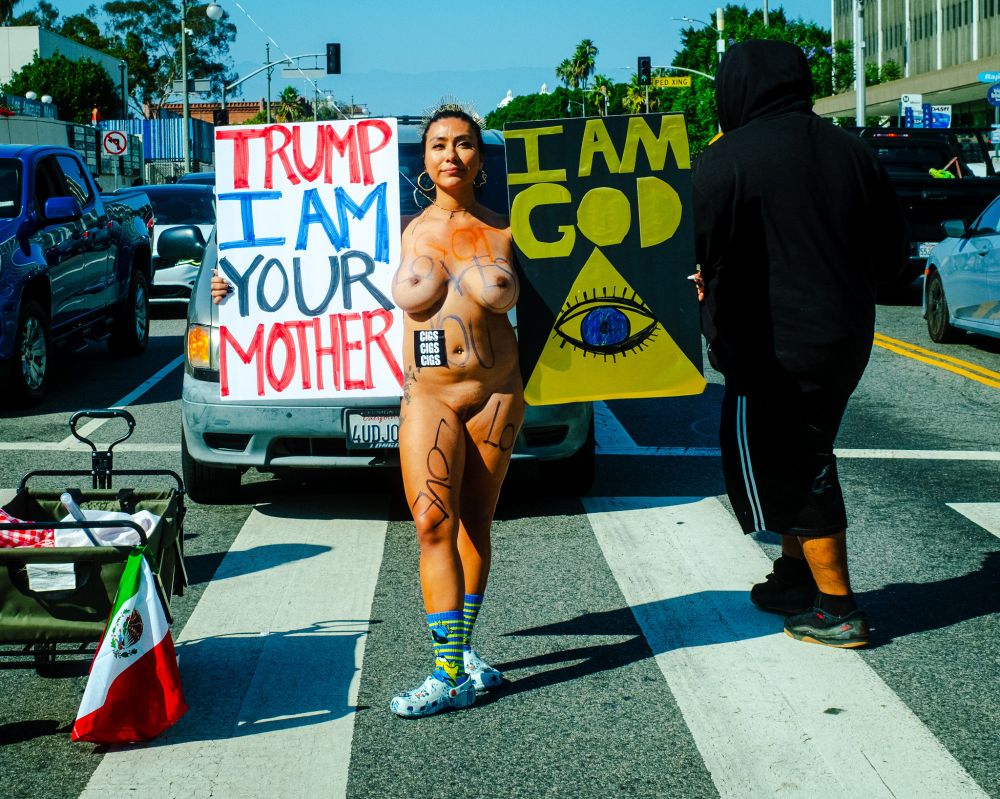 nude woman stands next to Mexican Flag in the middle of a cross walk, holds signs - one reading TRUMP I AM YOUR MOTHER and the other, showing cosmic eye in pyramid, reading I AM GOD 