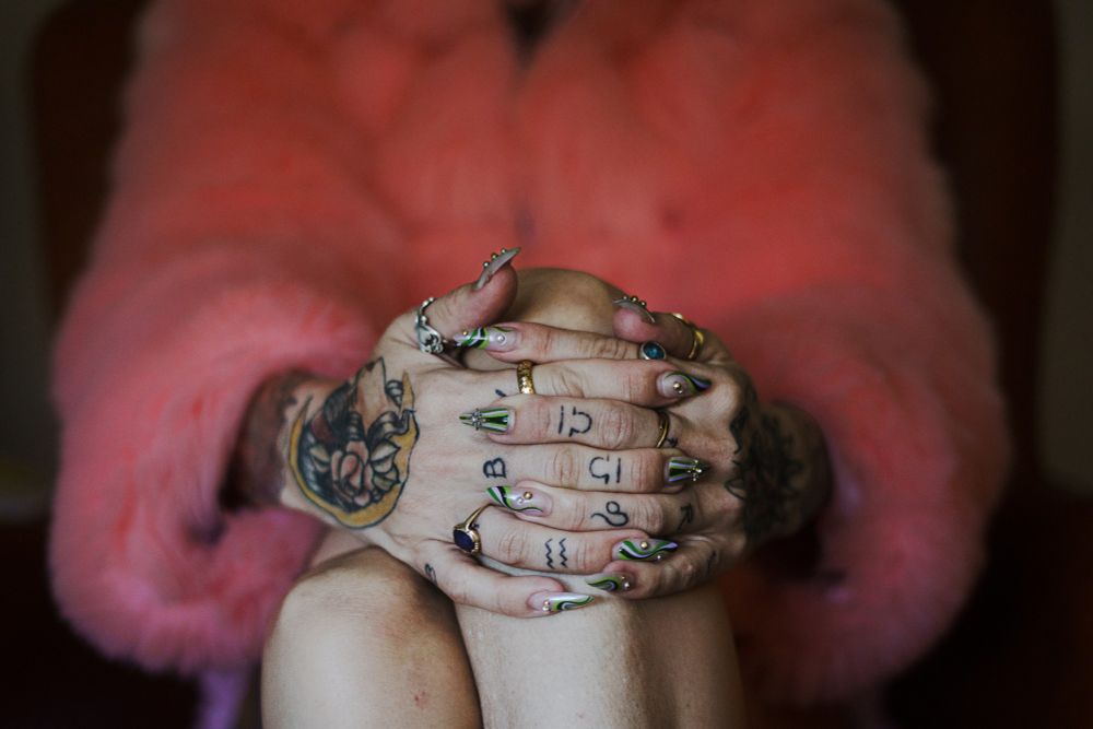 Kitt's tattooed hands folded together, nails, with her fuzzy pink coat in the background. 