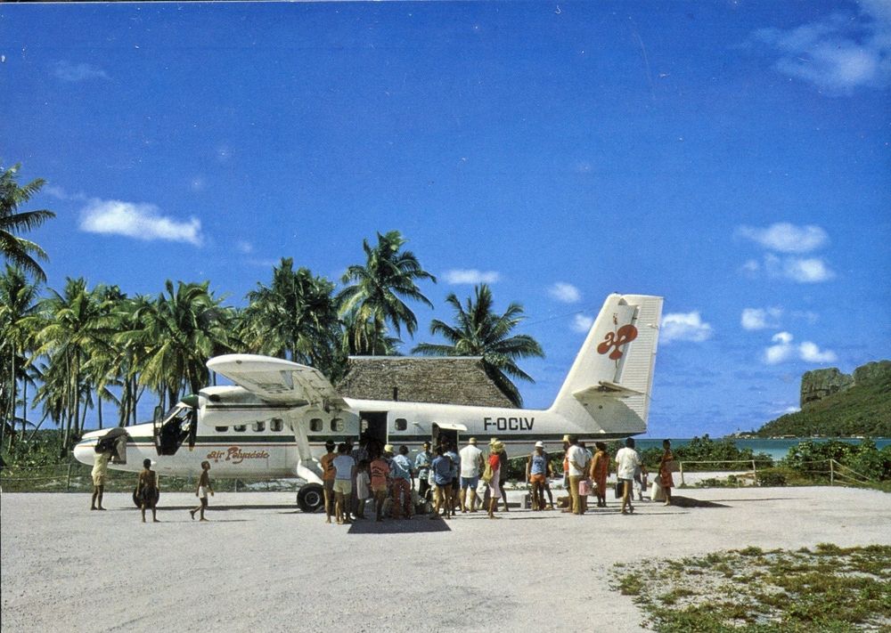 A de Havilland Canada DHC-6 Twin Otter of Air Polynésie sits on a sunlit island airstrip, framed by swaying palms and turquoise lagoon waters. Its cream-colored fuselage, marked F-OCLV, features the airline’s signature red tiare flower logo on the tail. A small crowd of passengers—some in bright island clothing, others in casual travel attire—gather at the open cabin door as they board or disembark. Children wander near the aircraft while adults wait in the warm, powdery coral sand that serves as the terminal ramp. Behind the plane, a thatched-roof hut blends seamlessly with the tropical setting, embodying the charm of remote Polynesian airfields. The sky is brilliantly blue, dotted with soft white clouds, and a steep, lush island peak rises in the distance. The scene captures the relaxed rhythm of inter-island travel in the South Pacific during the golden age of Air Polynésie, when the Twin Otter was the lifeline connecting scattered atolls and volcanic islands.