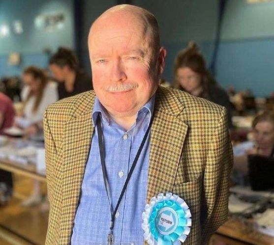 Reform councillor, bald-headed, moustachioed and wearing a yellowy-brown tweed jacket with a "Reform" rosette