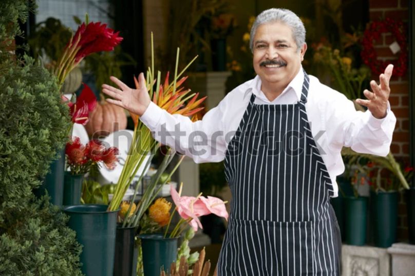 A watermarked stock photo of an older Italian gentleman in an apron, a shop owner of some kind, stepping up with a smile and his arms open in greeting. It’s the same picture every day. Today he is setting out flowers!