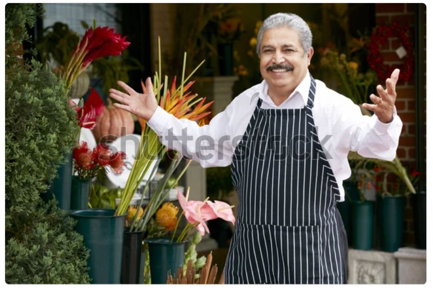 A watermarked stock photo of an older Italian gentleman in an apron, a shop owner of some kind, stepping up with a smile and his arms open in greeting. It’s the same picture every day. Today he is wearing fun shoes!