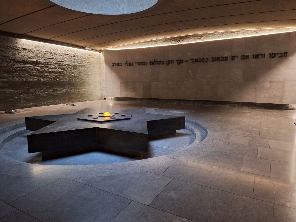 Ein schlichter Raum in Beigetönen. Am Boden ein schwarzer liegender Davidstern mit angedeudeter ewiger Flamme. An der Wand ein Zitat aus der Thora in mir unbekannter Sprache/Schrift