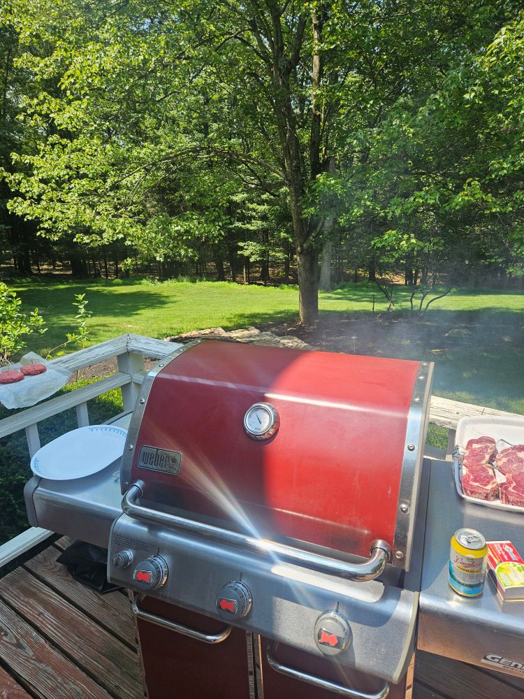 Grill with steaks, beer, and hamburgers nearby on a summer day.