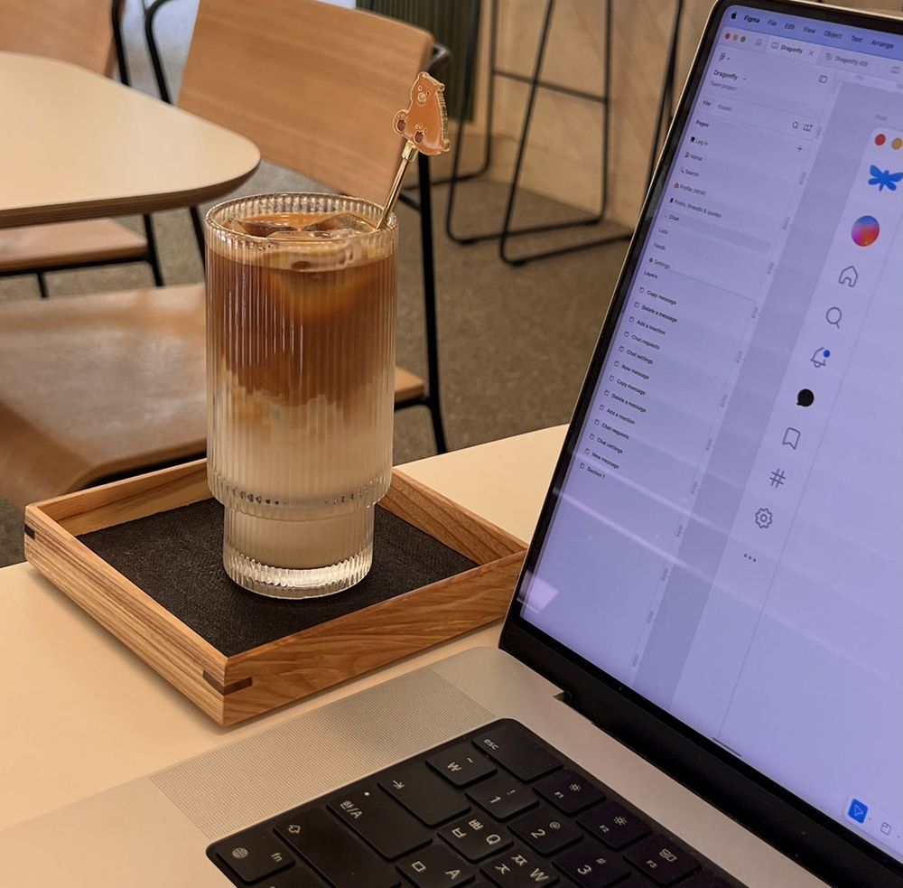 A glass of iced latte with milk and espresso layers sits on a wooden tray beside a laptop displaying a Figma design project. The café setting features modern wooden chairs and a clean, minimalist interior. A cute bear-shaped stirrer adds a playful touch to the drink.