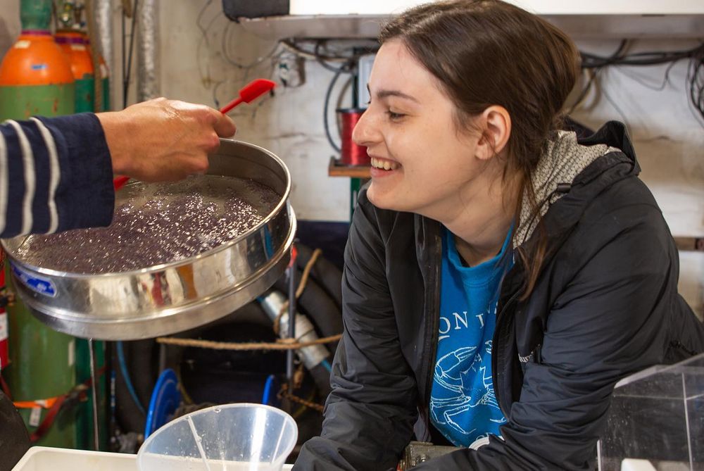 A scientist smiles while looking into a tray of brownish smushed zooplankton