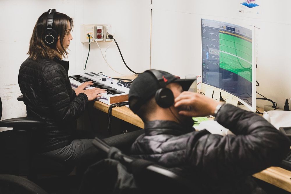 Two musicians wearing black jackets and headphones sit in front of a computer