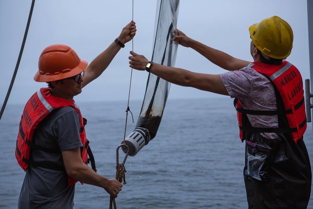 two scientists with life jackets and hard hats stand at the side of a boat holding a cone shaped net between them