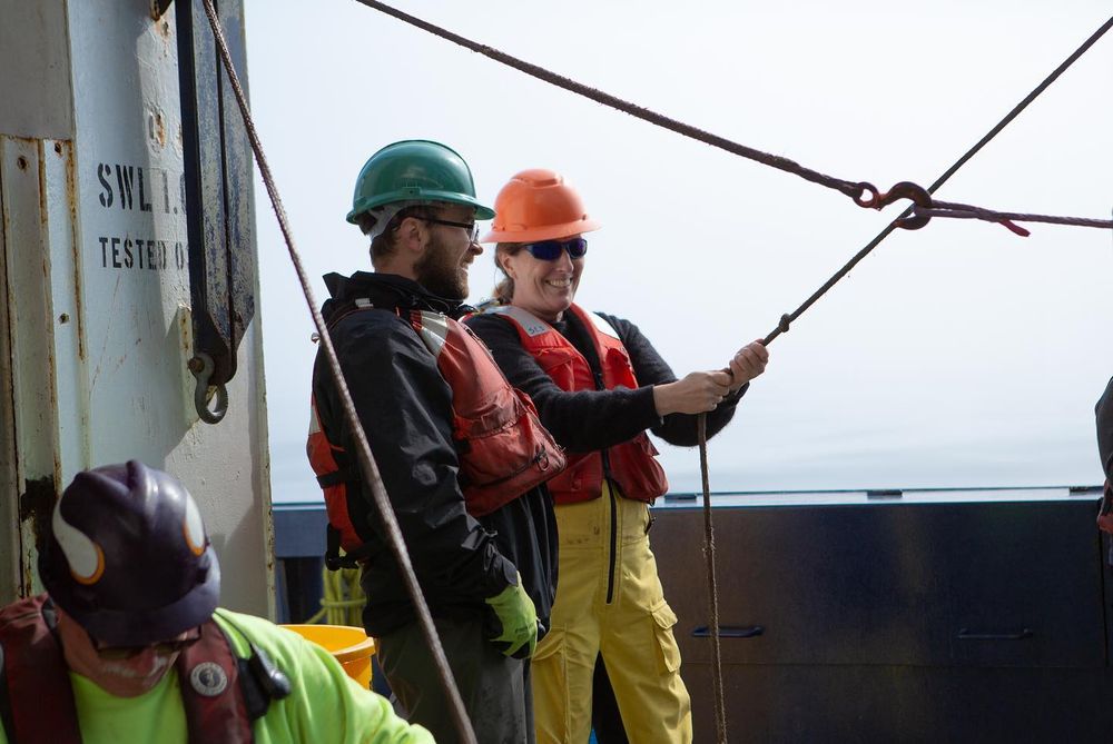two scientists wearing hardhats smile while one holds on to a cable