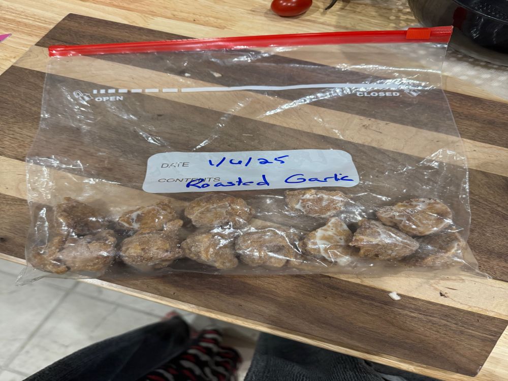 A gallon ziplock bag of frozen balls of roasted garlic on a striped wood cutting board. Bag has the date 1/06/25 and the words Roasted Garlic in blue sharpie. 