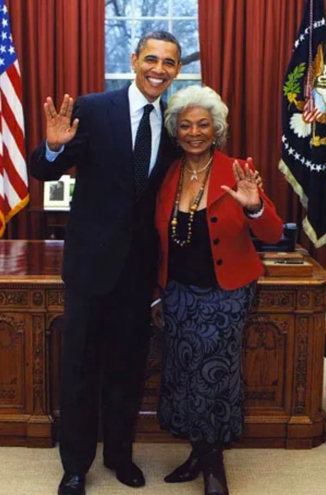 President Barack Obama stands in front of his desk in the Oval office with Nichelle Nichols. His arm is around her shoulders. He's wearing a dark blue suit, white shirt and black tie. She's wearing black boots, a purple skirt with a Paisley pattern, a black blouse, and a red jacket. They are both smiling and giving the Vulcan salute 🖖🏼