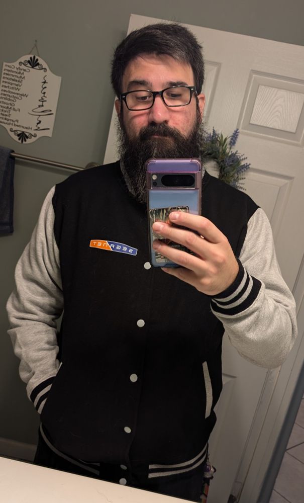 A bearded Midwestern white dude wearing a black and grey SegaNet varsity jacket while taking a millennial flashless bathroom mirror selfie with a blue Pixel smartphone and kind of making that stupid bear face