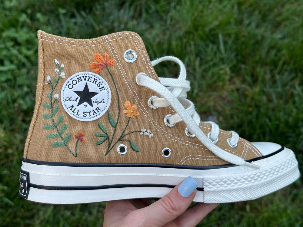A light brown converse chuck Taylor embroidered with flowers in earth tones surrounding the logo. 