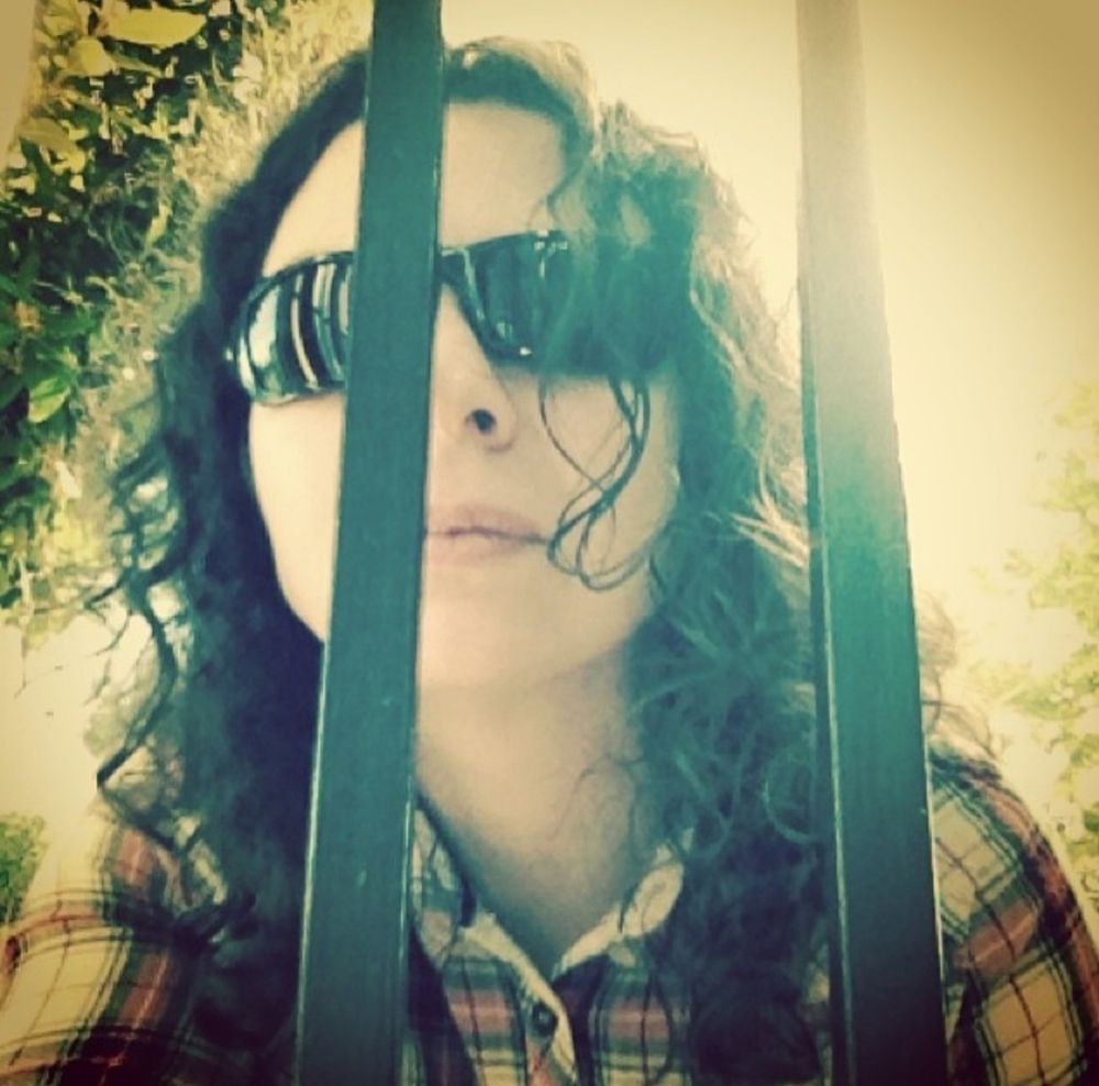 Yo con gafas de sol, pelo largo y camisa de cuadros mirando desde fuera a través de las rejas de una ventana. 