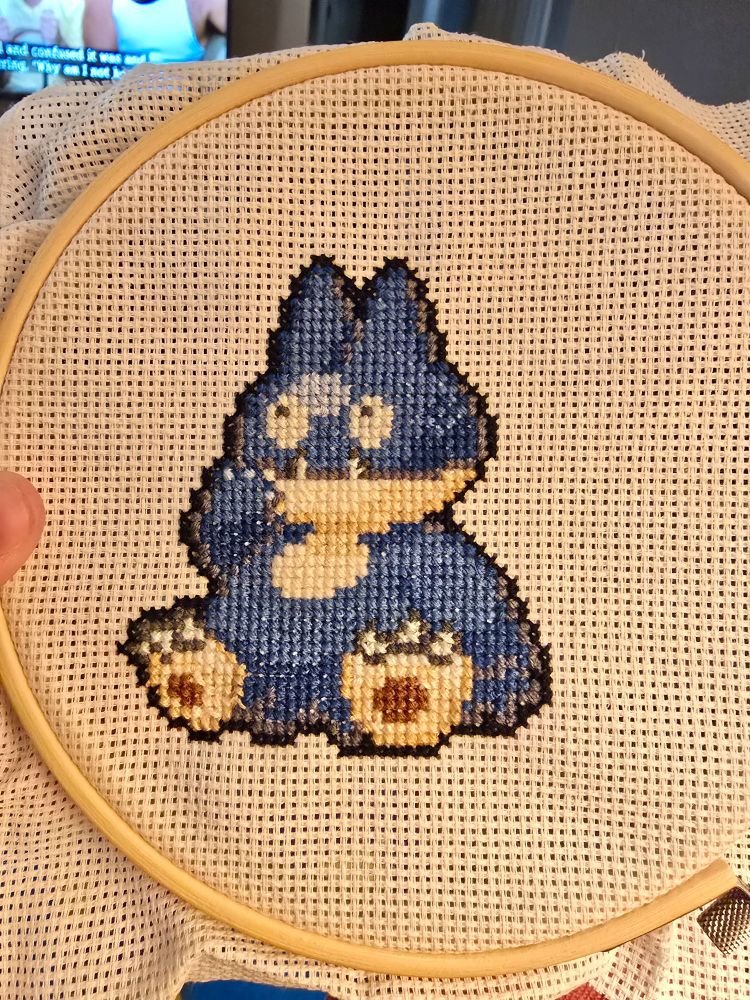 A munchlax sits with it's paw close to its mouth. It is a pixilated rendered as a crossstitch still on the hoop. 
