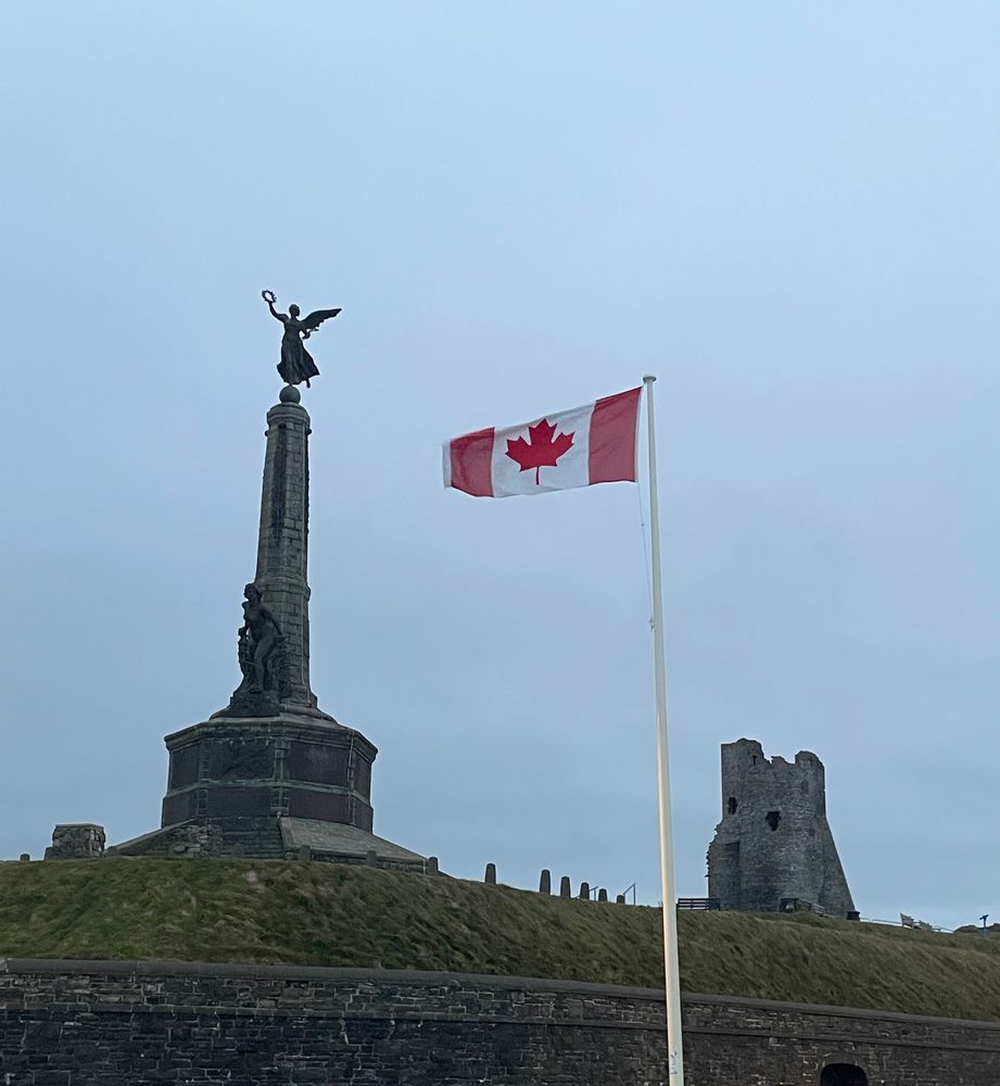 On a grey-blue sky background, we see on the left a statue of a winged woman on a tall pedestal, on the right the ruin of a castle turret, and in the middle a Canadian flag on a tall flag post in the foreground. Between the monuments and the flag post, there is grass, and a stone wall.
