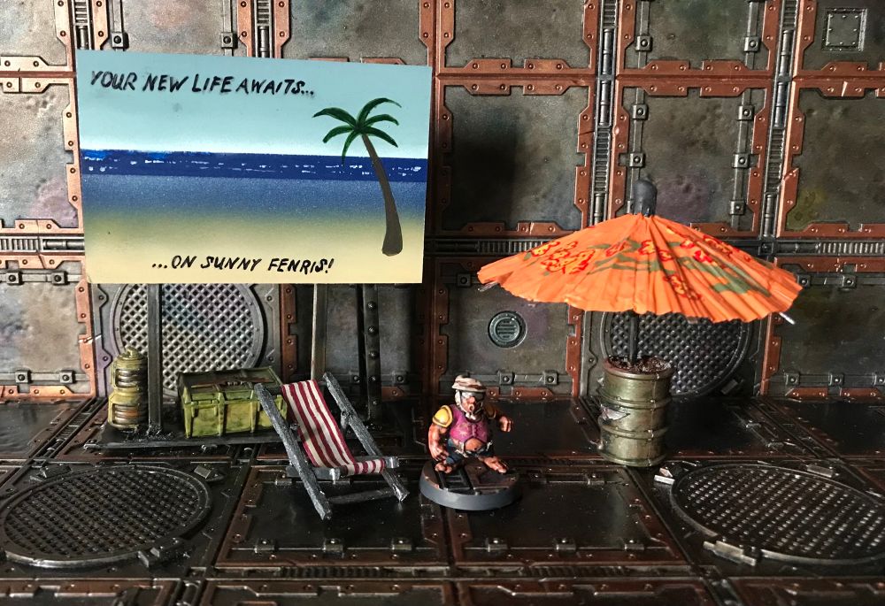 A Ratling in a Hawaiian shirt stands amidst a deck chair, a parasol and a bill board advertising holidays.