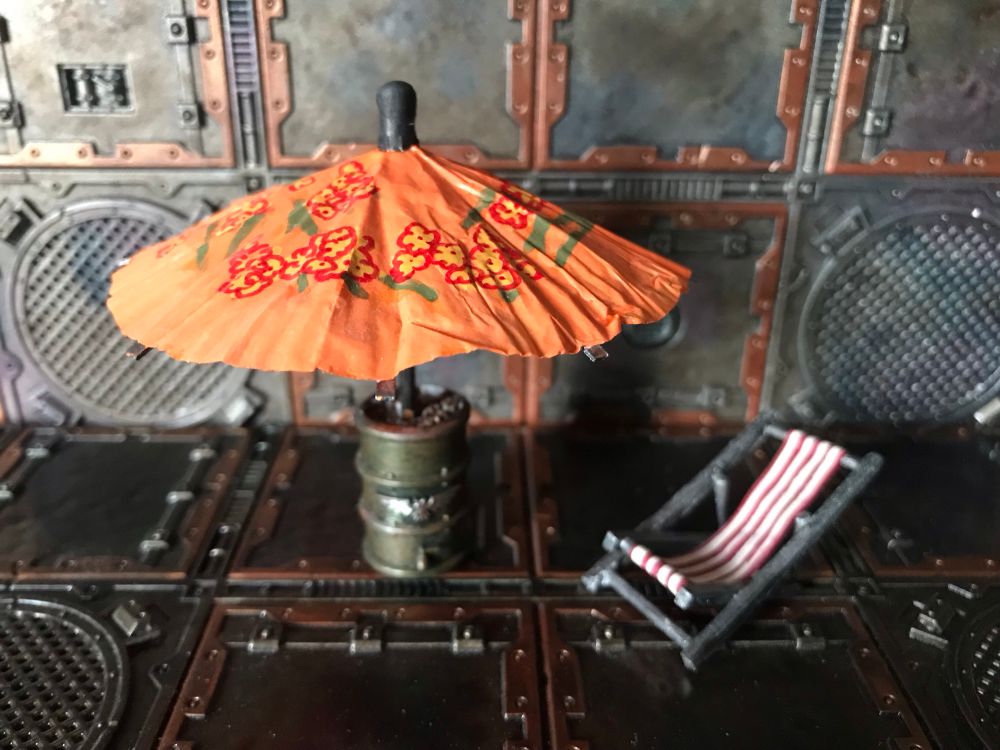 A parasol made from a cocktail umbrella and a plastic GW barrel.  Next to it is a deckchair made from match sticks and paper.