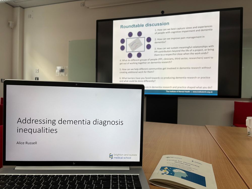 A photo taken from the perspective of someone attending a conference. There is a laptop set up in front of the camera, with the title slide 'Addressing dementia diagnosis inequalities' on its screen. It sits on a glossy wooden table, which also has a booklet for the conference sitting next to the laptop. In the background is a large presentation screen with a slide suggesting questions for a roundtable discussion.
