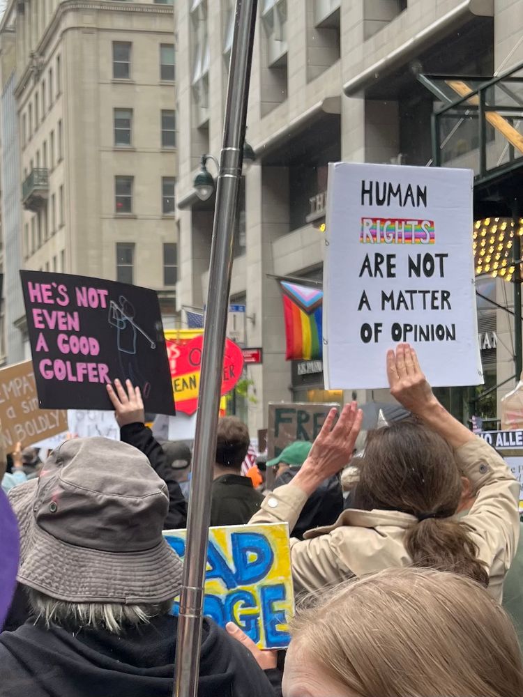 Two handwritten signs
1. Pink lettering on black paper saying “He’s not even a good golfer”
2 “Human rights are not a matter of opinion” (with the Rights in white lettering on a rainbow block)