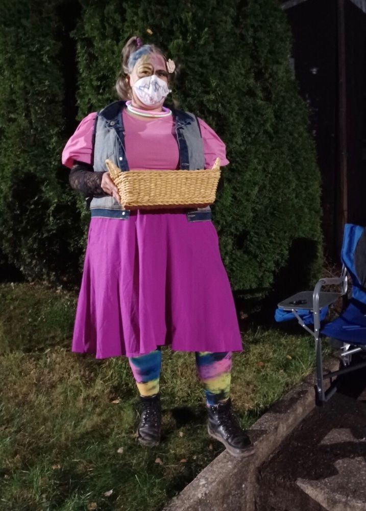 Photo of me dressed as Weird Barbie, standing on the sidewalk in front of my house at night and holding a basket of candy. I'm wearing a pink shirt with puffy sleeves, an inside-out denim vest, a pink skirt, multi-colored tights, black boots, glow-stick necklaces, and a duckbill N95 mask. My face and mask are scribbled on with various colors, and my hair is in several messy ponytails/braids sticking out from my head.