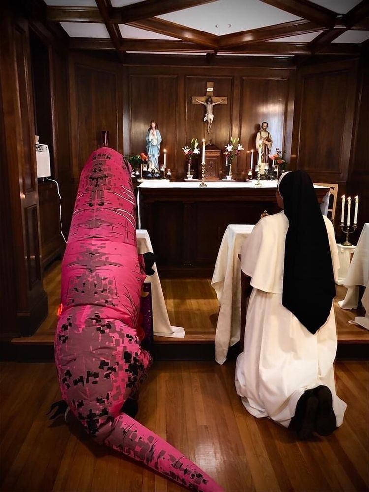 Photo of a person in a pink inflatable T-rex costume and a nun in a habit kneeling side-by-side in front of the alter in a chapel