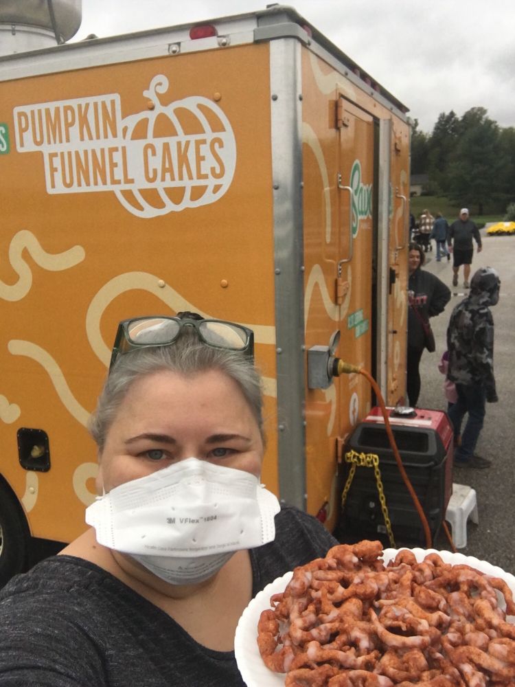Selfie of me holding a pumpkin funnel cake up to the camera. Behind me is a yellow food truck with "PUMPKIN FUNNEL CAKES" written on the side. I am wearing a grey shirt and a white V-Flex N95 mask.