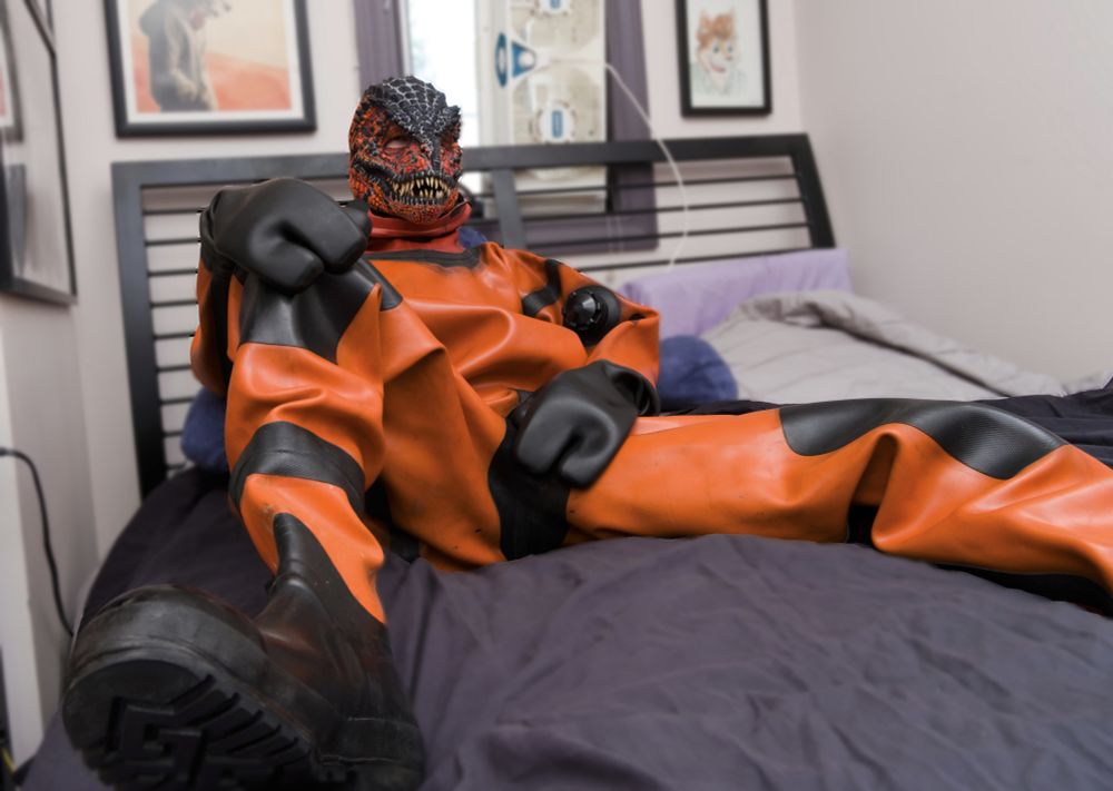 snickers? sitting on the bed with his legs spread open wearing an orange drysuit, diving mitts, and an orange t-rex mask