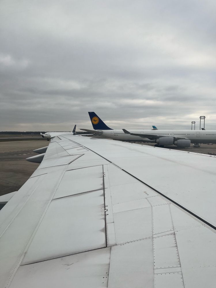 Sicht aus einem Flugzeugfenster auf eine Tragfläche. Man sieht graue Wolken am Himmel und ein anderes Lufthansaflugzeug.