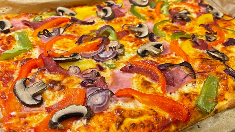 A rectangular homemade pizza after baking, topped with a colorful mix of ingredients. Visible are slices of salami and ham, red, yellow and green bell peppers, red onion, and sliced mushrooms. The pizza is laid on parchment paper in a baking tray, with a tomato sauce base and baked mozzarella and gouda cheese underneath the toppings.