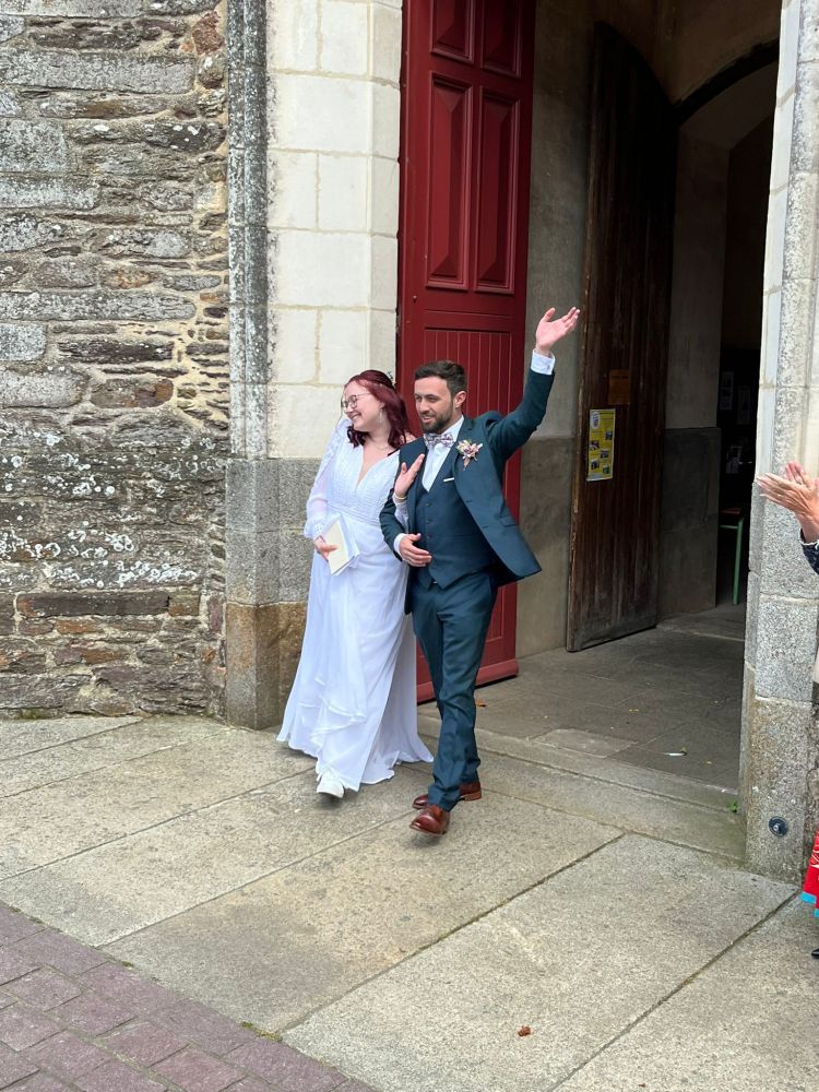 Anaïs et Enzo marchent en sortant de l'église où ils viennent de se marier. Enzo, en costume vert foncé, lève la main gauche en signe de joie. Anaïs, en robe blanche, sourit et regarde les invités.