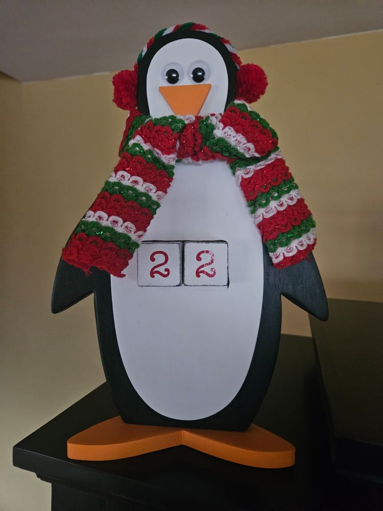 A Christmas countdown calendar in the shape of a penguin with a red, white and green scarf, candy cane earmuffs, and googly eyes.
