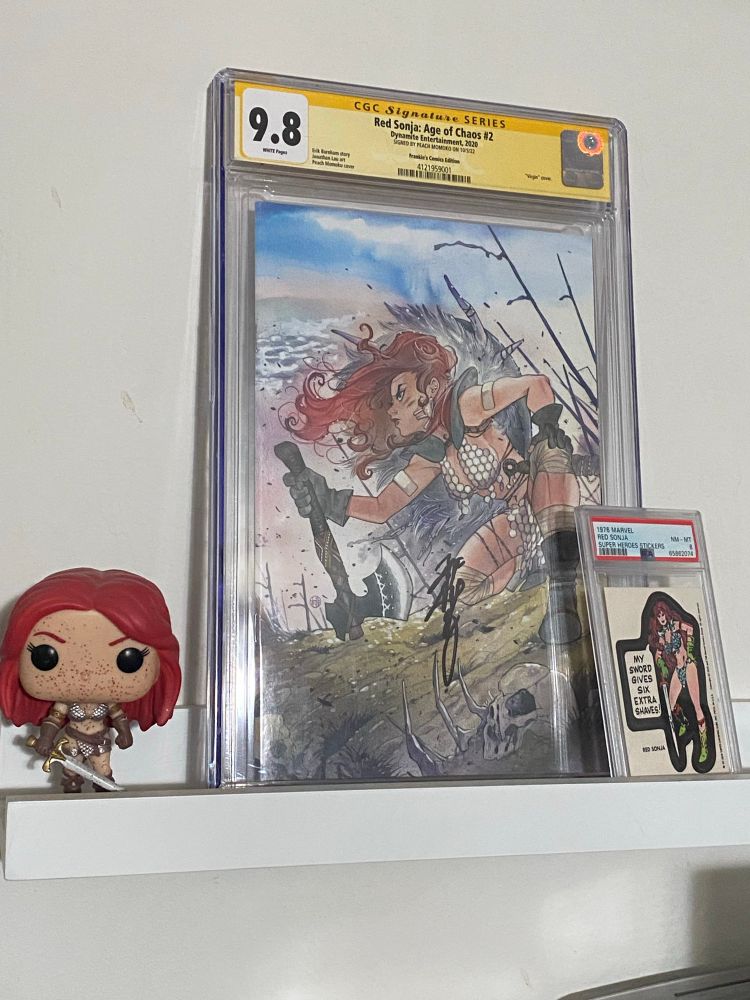 Alt text: Photograph of some comic-related things on a small shelf. First, there is a Red Sonja Funko Pop (blood spattered version). There is a PSA-graded Red Sonja sticker from the 70s in which she is pictured leaning on her sword and saying “my sword gives six extra shaves!” I have no idea what that is supposed to mean. And finally there is a CGC-graded copy of Red Sonja: Age of Chaos 2, the cover of which has a beautiful Peach Momoko painting of Red Sonja crouched on a hillside. She is holding the haft of an axe, its head on the ground in front of her. She is wearing her traditional scale mail bikini armor and her red hair is flowing in a breeze coming from behind. There are tall spears protruding at a slight angle in the background and in the foreground there are bones in the grass.