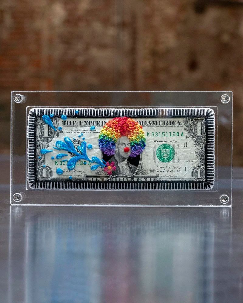 A one dollar bill in a clear frame with George Washington embellished with embroidery, giving him a rainbow clown wig and novelty flower badge squirting water.