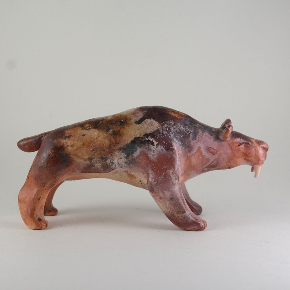 A photo of a ceramic saber tooth cat sculpture on a white background. The sculpture has swirling colors of orange, tan, burgundy, pink, and black.