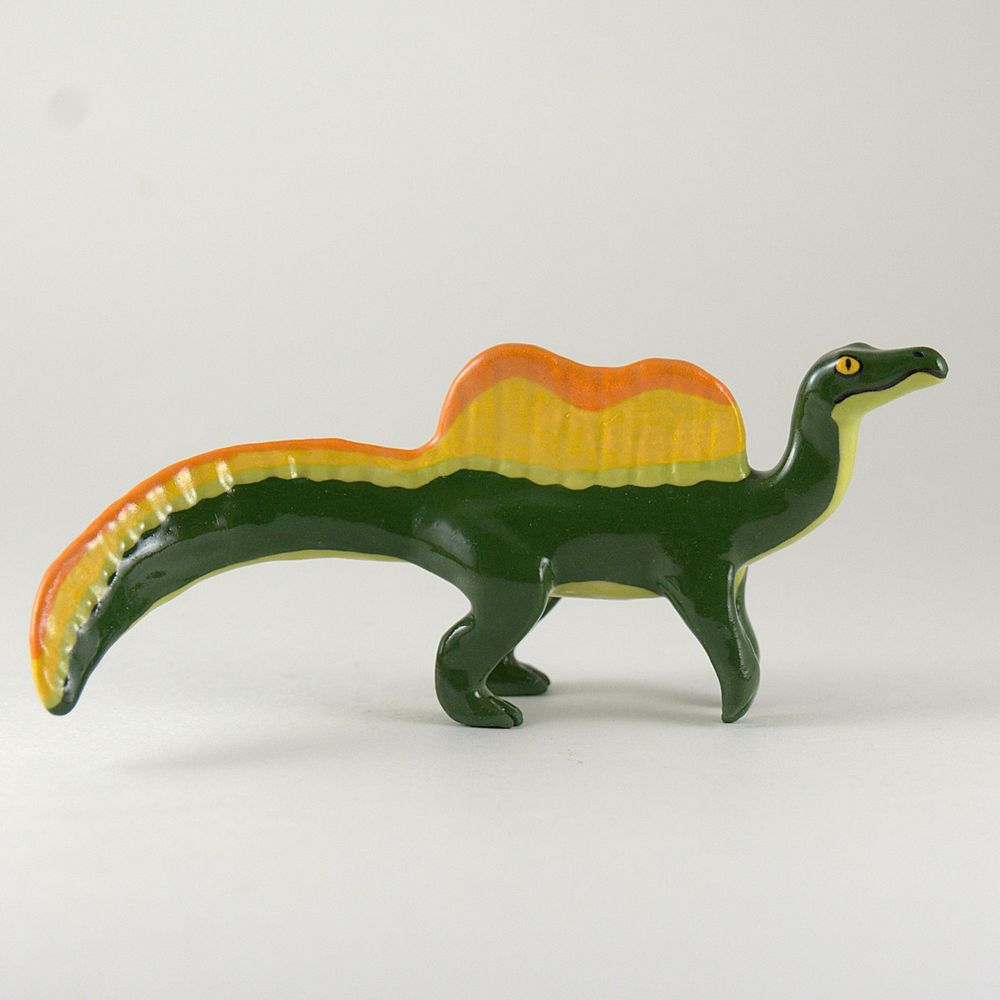 A photo of a ceramic spinosaurus on a white background. The sculpture is mostly green with yellow and orange on the tail and dorsal sail.