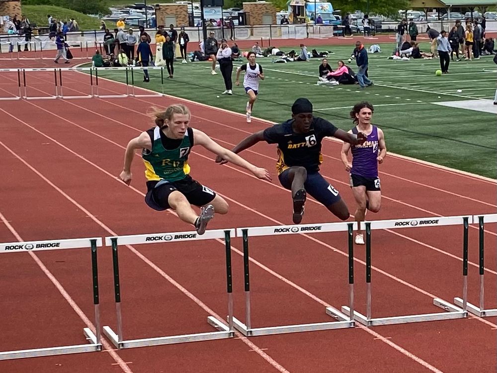 The same competitor in the same green and yellow jersey vaults a hurdle in a virtual tie with the computer in the next lane. Two other competitors are slightly behind them on the track.   