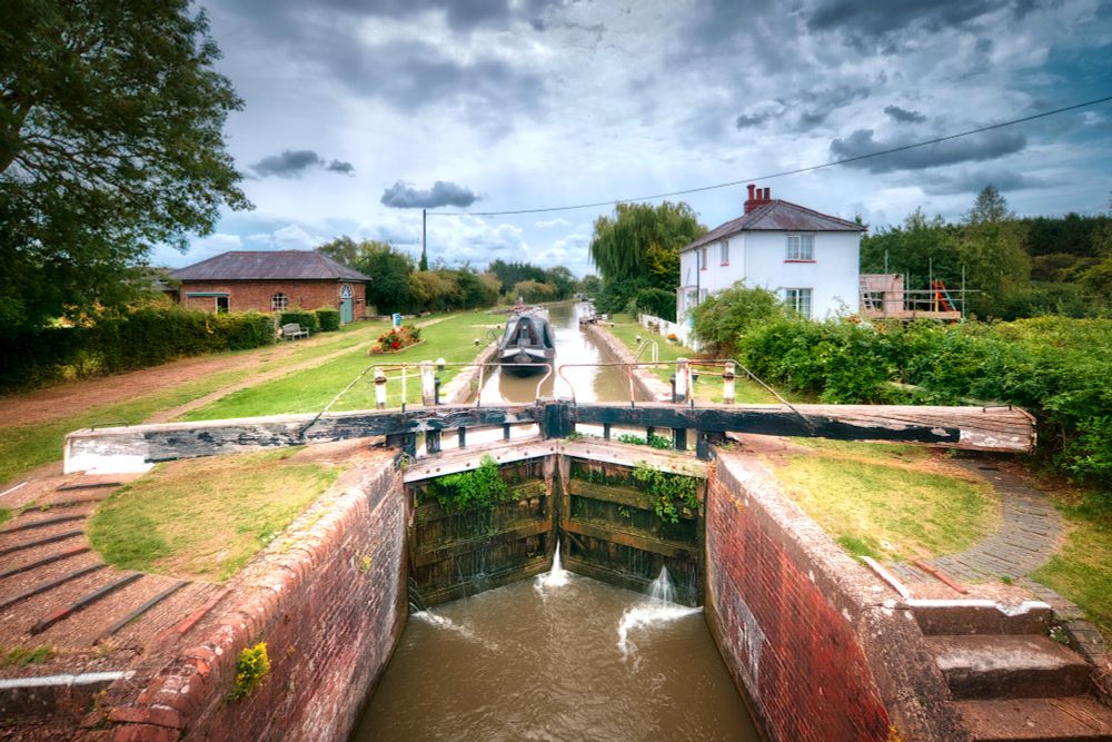 Photo of a narrowboat entering a wide lock taken from the bridge that spans the canal below the lock. Looking on to the lock to the right stands the two-storey, white, former lock-keeper's cottage. A wider expanse to the left of the lock between lock and towpath is where a second narrow lock once was but is now filled in. On the other side of the towpath stands the single-storey brick-built former pumping station.