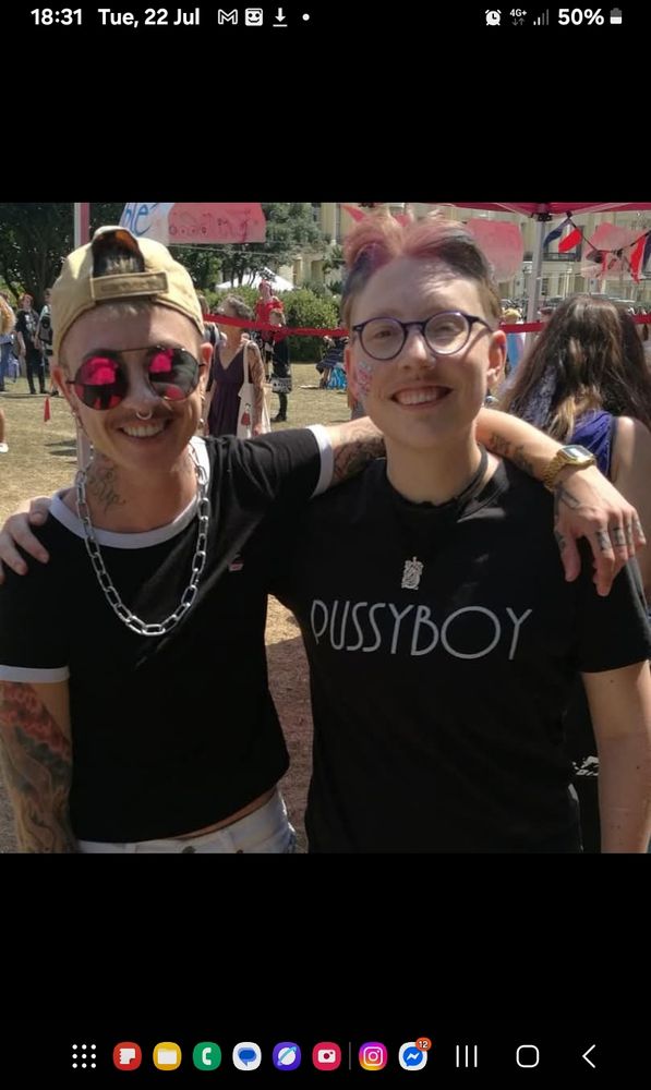 A photo of two transgender men. They are both smiling. The person on the left is wearing a backwards cap, sunglasses, a chunky chain necklace and a black t-shirt. He has his arm around the other person's shoulders. The person on right is wearing glasses, has a transgender flag painted on their face and is wearing a black t-shirt with the word fussy boy on it. 