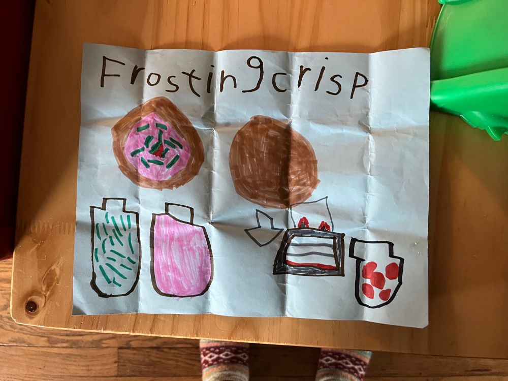 Light blue paper with the heading Frosting Crisp. A drawing of the stages of making a cookie begins at top right with a large round brown cookie. With an arrow underneath leading to the drawing of an oven. Moves to left top with the brown cookie covered in pink frosting and rainbow sprinkles with a cherry in the middle. Below the iced cookie are drawings of two jars one with sprinkles and one with pink frosting. There is a drawing of a jar next to the oven with cherries in it (because 5 forgot to include it initially-LOL). 