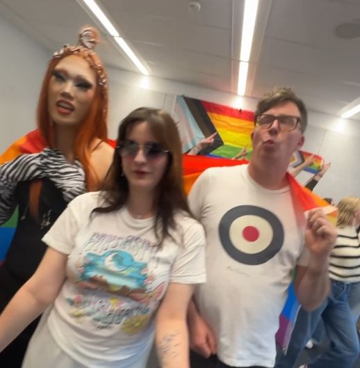 A photo of three people: On the left a dragqueen with orange hair in a black dress, in the middle a person with sunglasses, brown hair and a white t-shirt, on the right a person with short brown hair, glasses, and a white shirt with circles on it. They carry a rainbow flag. In the background more people are somewhat visible, they seem to be dancing. On the wall in the background, there is a progressive pride flag.