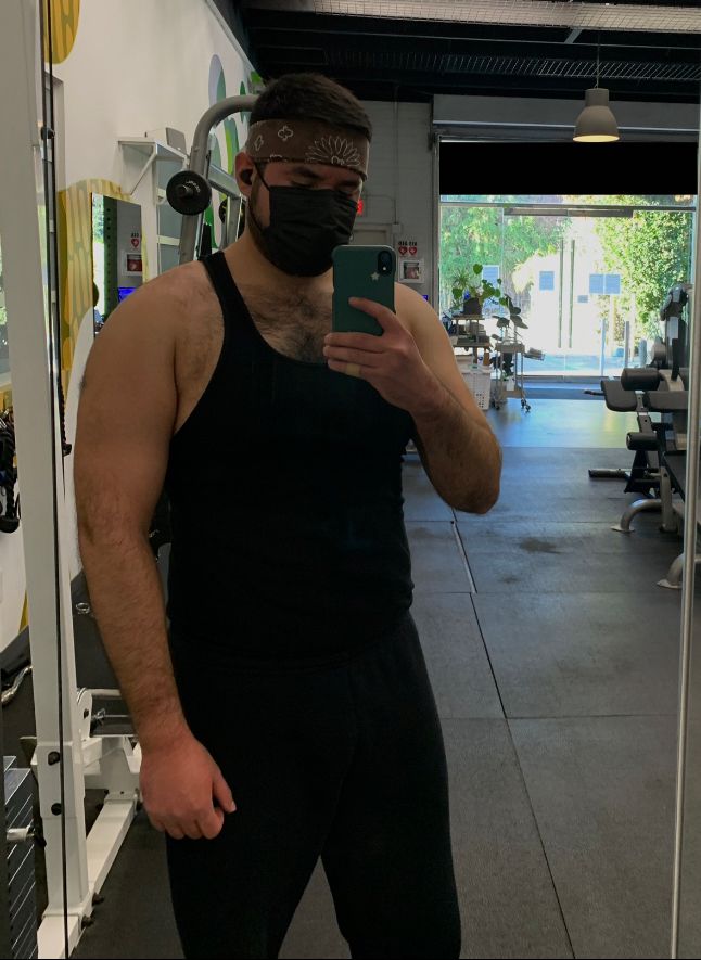 A mirror selfie at the gym. I’m wearing black sweatpants, a black tank top, a black mask, and a brown paisley bandana as a sweatband. There is visible gym equipment behind me. 