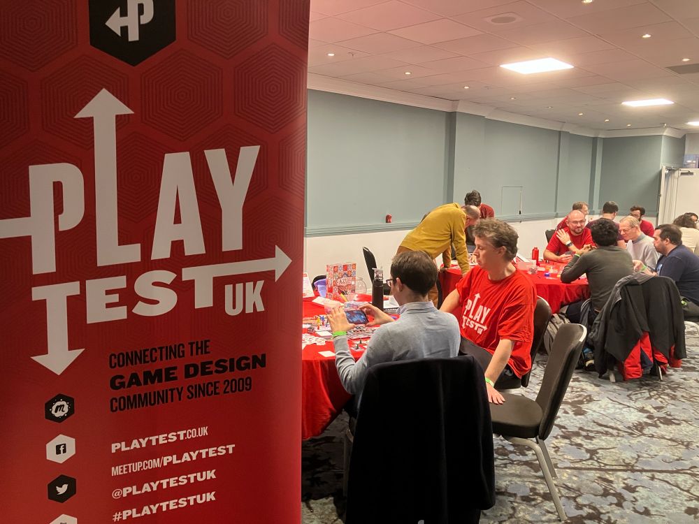 Designers and playtesters playtesting game prototypes with the Playtest UK banner in the foreground. 