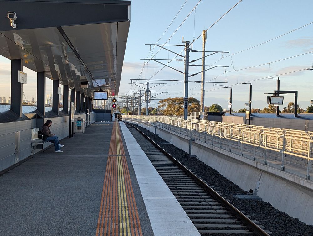 Auto-generated description: A person sits on a bench at an empty outdoor train station platform under a clear sky with train tracks extending into the distance.