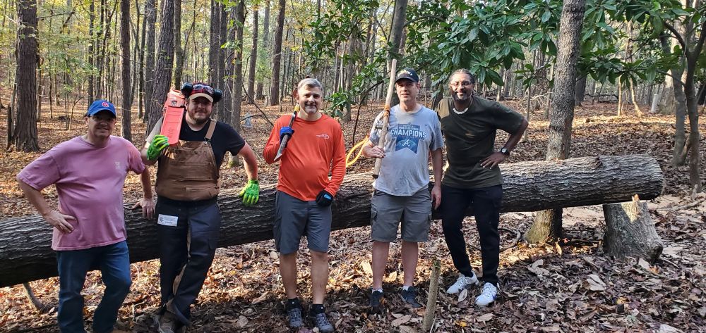 Five volunteers standing alongside a fallen tree in a wooded historic cemetery, holding forestry tools