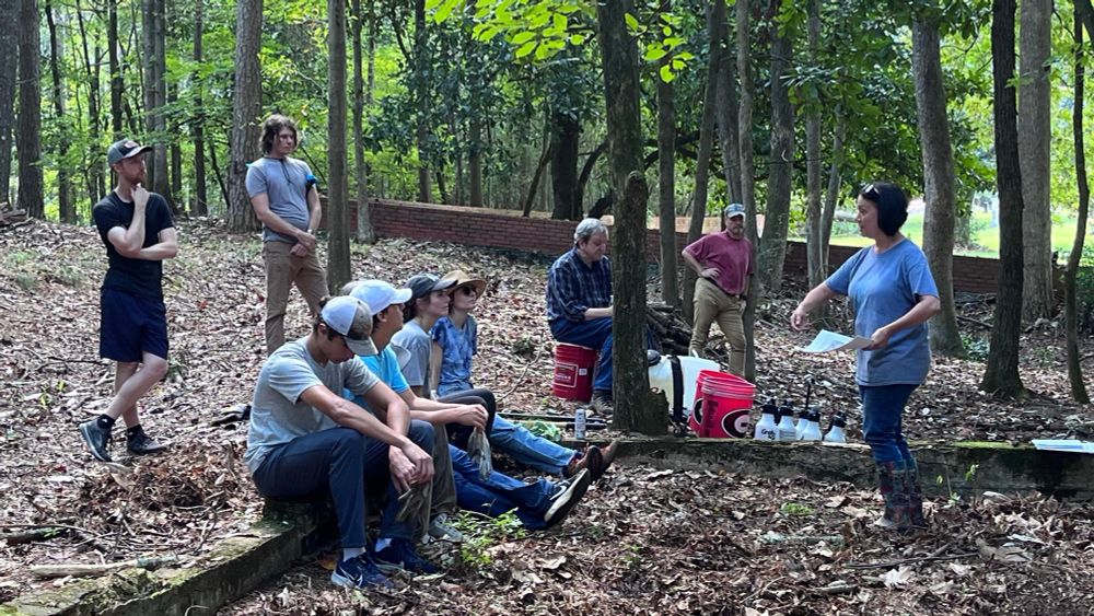 8 volunteers sitting in a wooded cemetery listening to a presentation about headstone cleaning