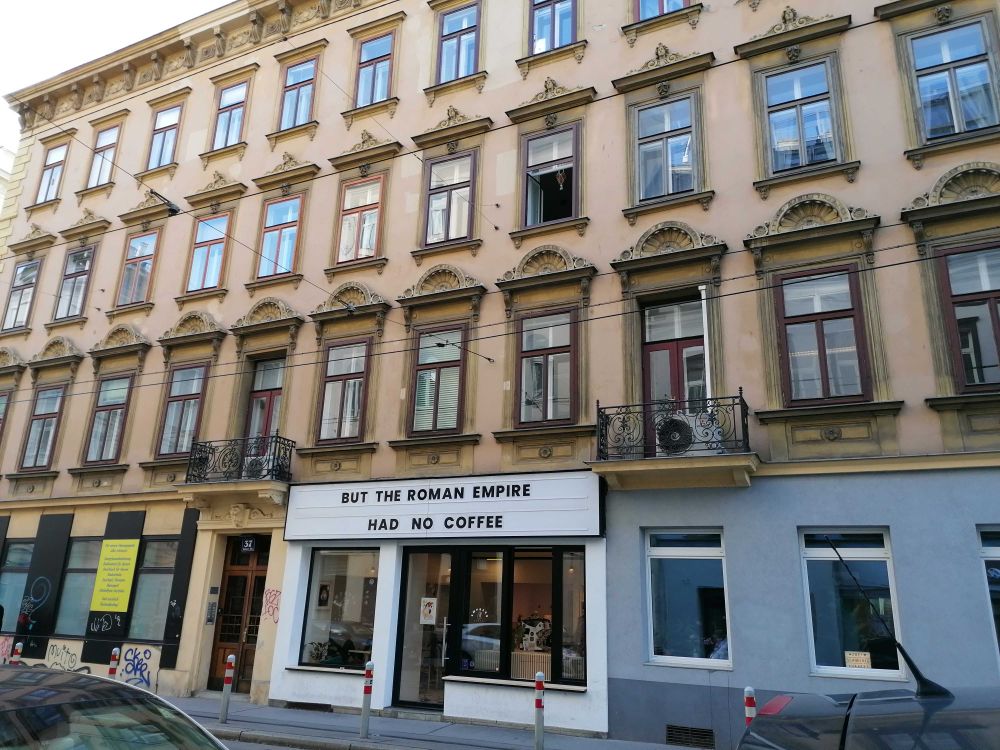 A picture of a small café, think hole in the wall. Its sign is one where the letters can be changed, like at an old movie theater. They frequently change it. It's often a meme or a song lyric. This time the sign says "But the Roman Empire had no coffee". It's a reference to the "thinking of the Roman Empire" meme that was going around two weeks or so ago.