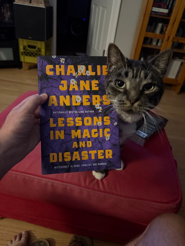 A copy of the hardcover of LESSONS IN MAGIC AND DISASTER by Charlie Jane Anders, with Mr. Darcy, a monster cat, peeking around from behind the book.  If you don’t find this adorable, you may be a monster too…