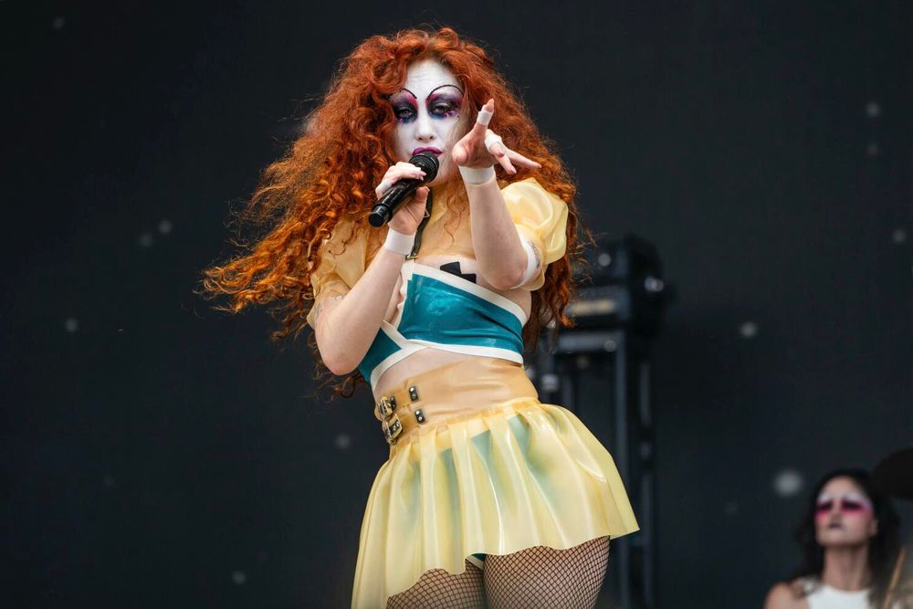 foto da Chappell Roan em cima do palco num show com uma maquiagem com base bem branca, cabelo cacheado bem ruivo/vermelho e esvoaçante. a cantora veste um topzinho que parece um biquíni verde e uma saia de látex amarela 