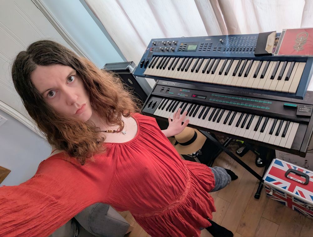 Trans girl posing in front of a few Yamaha Synthesisers. 